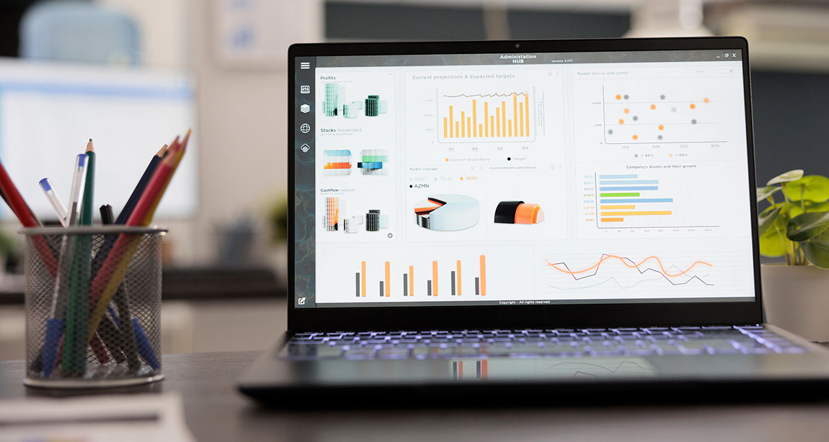 An open laptop on an office desk showing a dashboard of bar, pie and line charts, beside a cup of pens and a potted plant