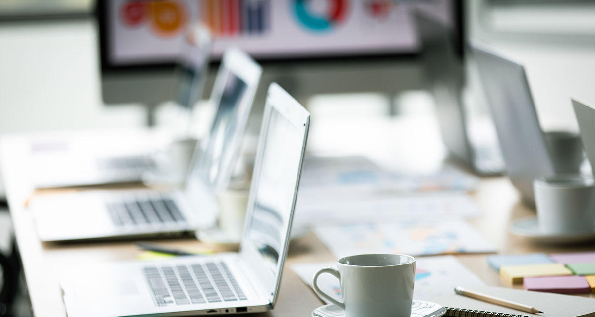 Open laptops, coffee cups, sticky notes and printed charts on an empty conference table, with a sales performance dashboard on a screen behind