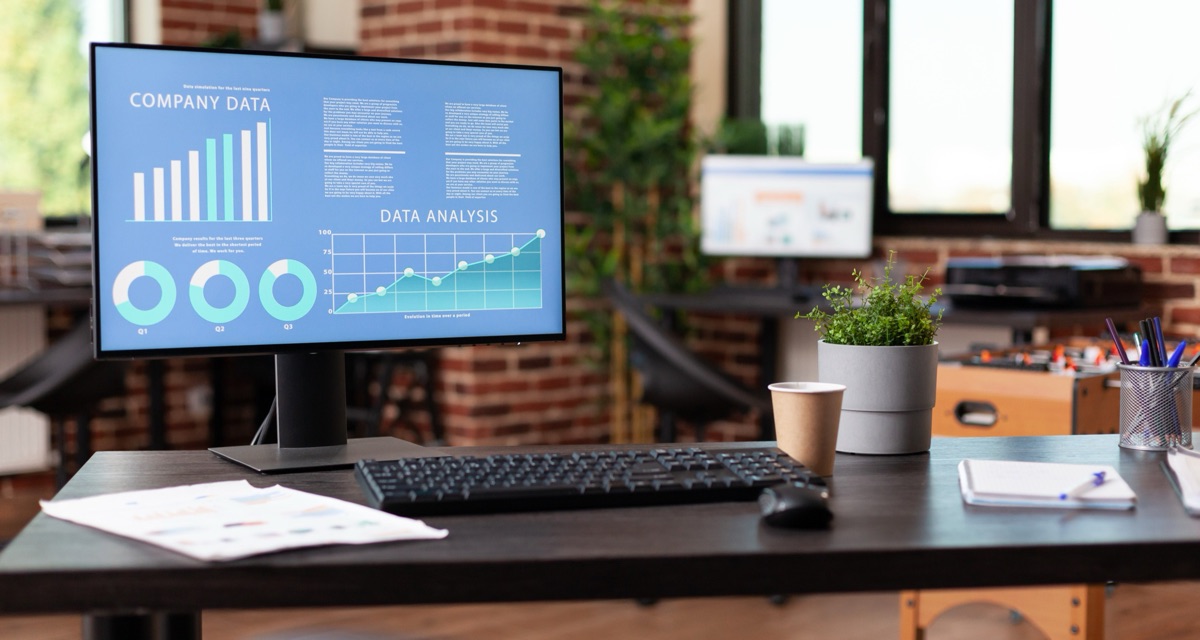 A desk monitor showing company data bar charts, quarterly donut charts and a rising line chart, beside a keyboard, printed reports and a coffee cup in a bright office