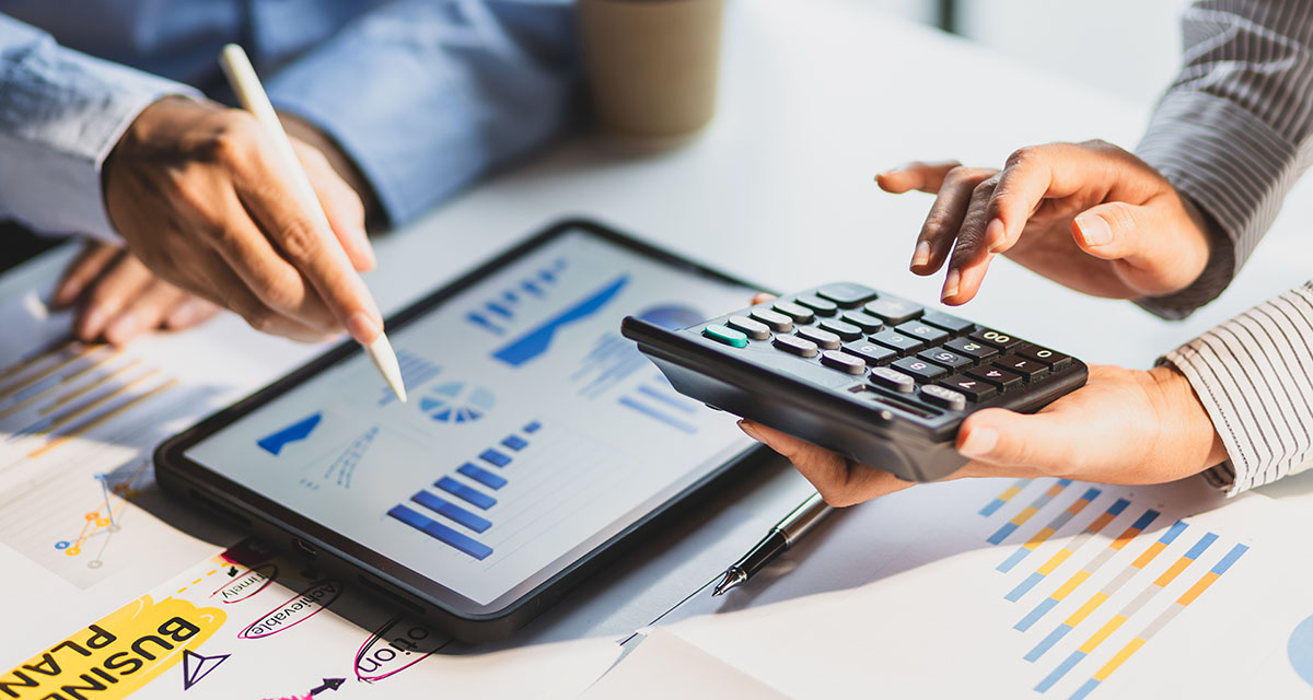 Two colleagues reviewing budget charts on a tablet while one works a calculator over printed financial reports
